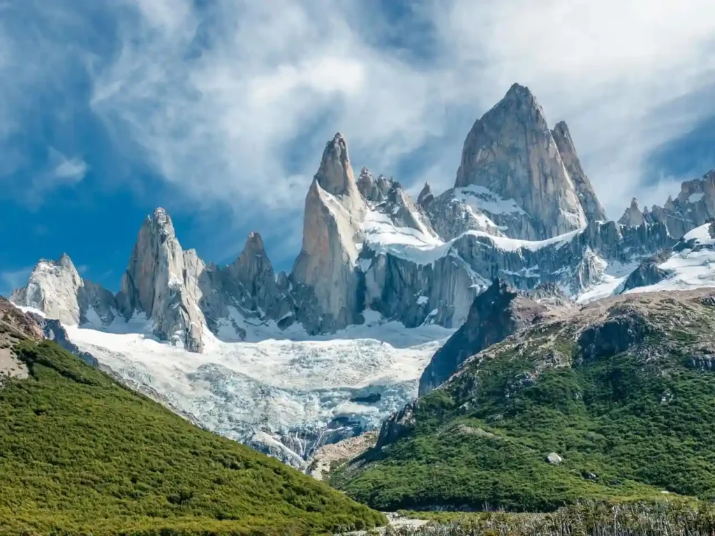 The jagged, snow-capped peaks of Mount Fitz Roy rise sharply behind a glacier and green foothills, under a bright blue sky with wispy clouds.