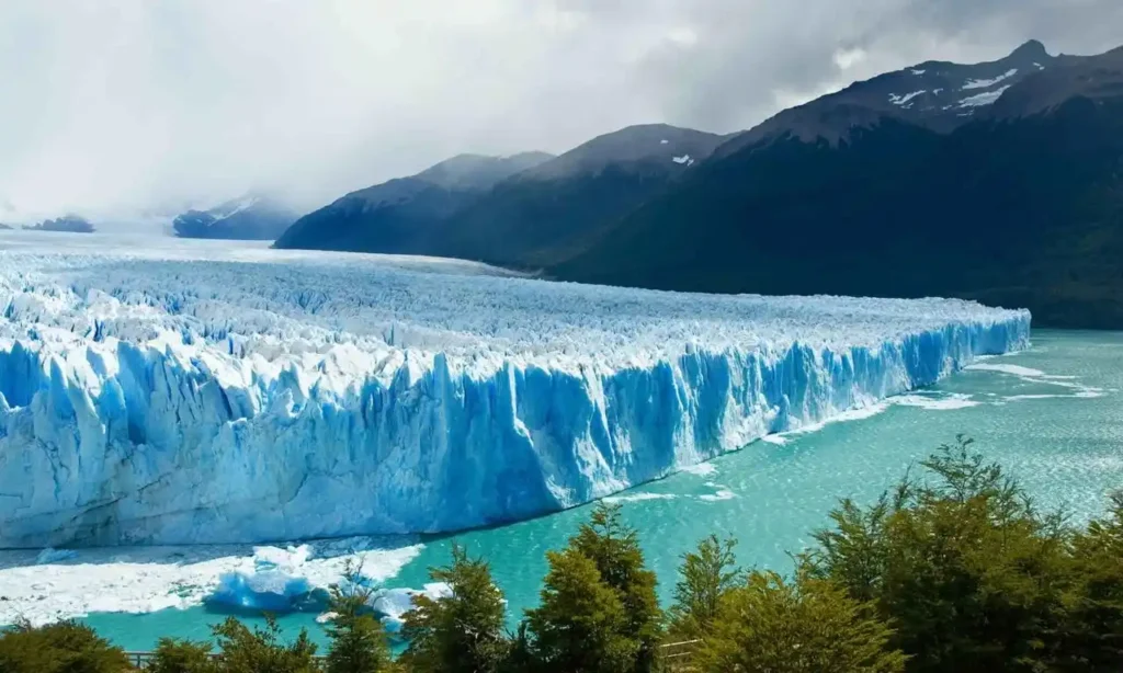 A massive, blue-tinged glacier with a jagged face descends into a turquoise lake, with a backdrop of forested mountains under a cloudy sky.