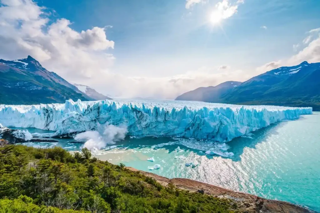 The vast, blue face of the Perito Moreno Glacier spans a turquoise lake, with a large chunk of ice calving into the water, under a bright sun and partly cloudy sky.