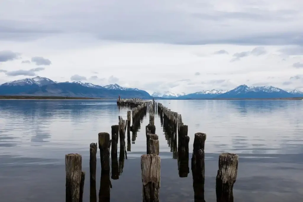 A line of old, weathered wooden pier posts extends into a calm, reflective lake with snow-capped mountains in the distance under a cloudy sky.