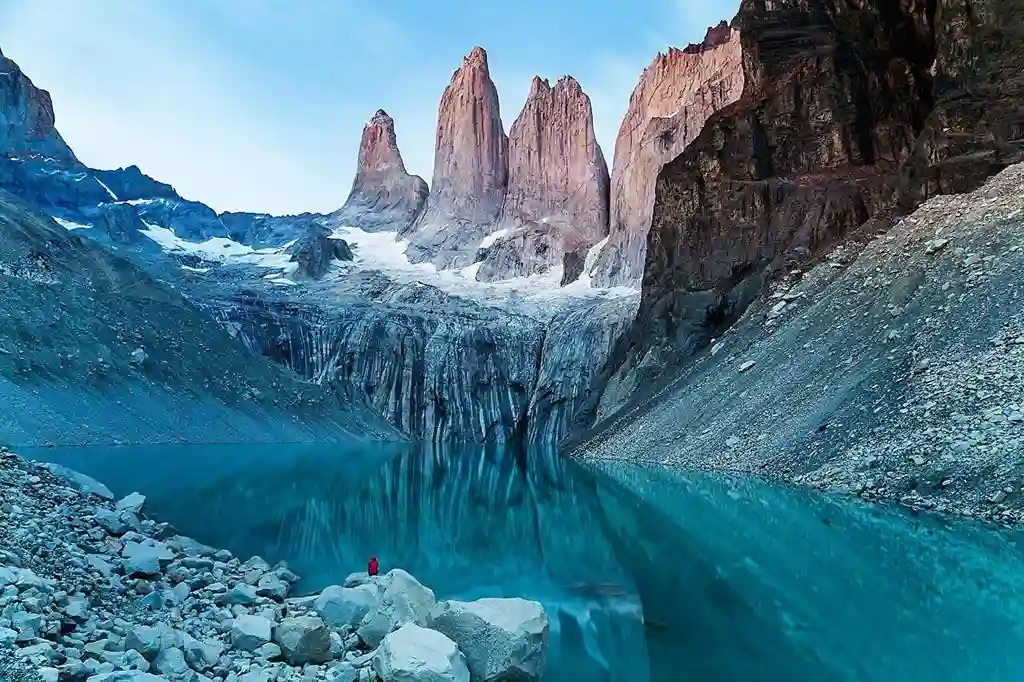 Three colossal granite towers rise from behind a glacier and a vivid turquoise lake, with a single person in a red jacket standing on the rocky shore.