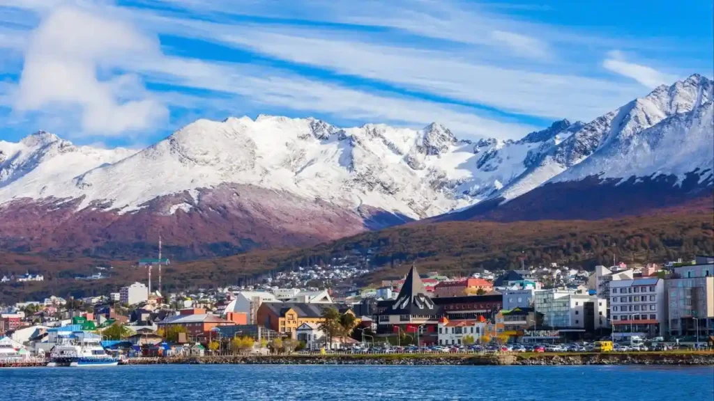 The colorful city of Ushuaia is nestled on the shore of a bay, with a dramatic backdrop of snow-capped mountains under a blue, cloudy sky.