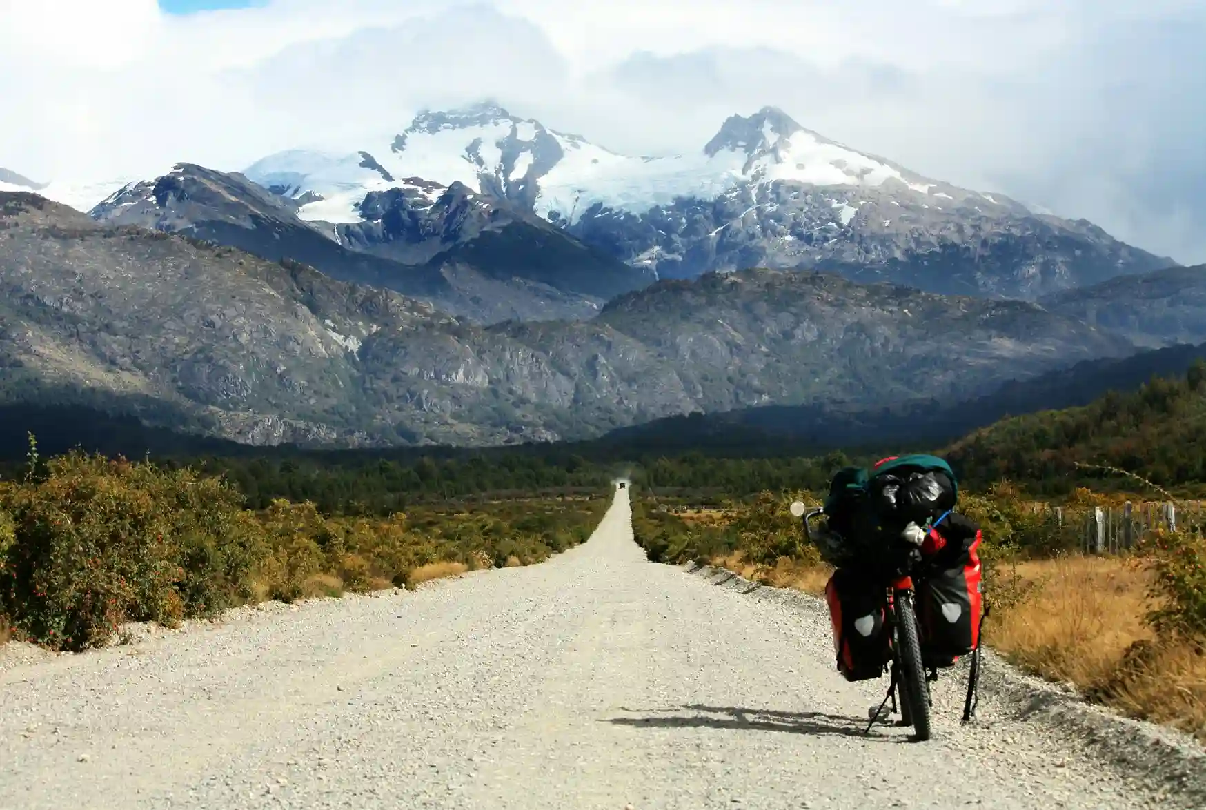 A mountain bike with saddlebags parked on a long, straight gravel road with snow-capped mountains and green fields in the background.