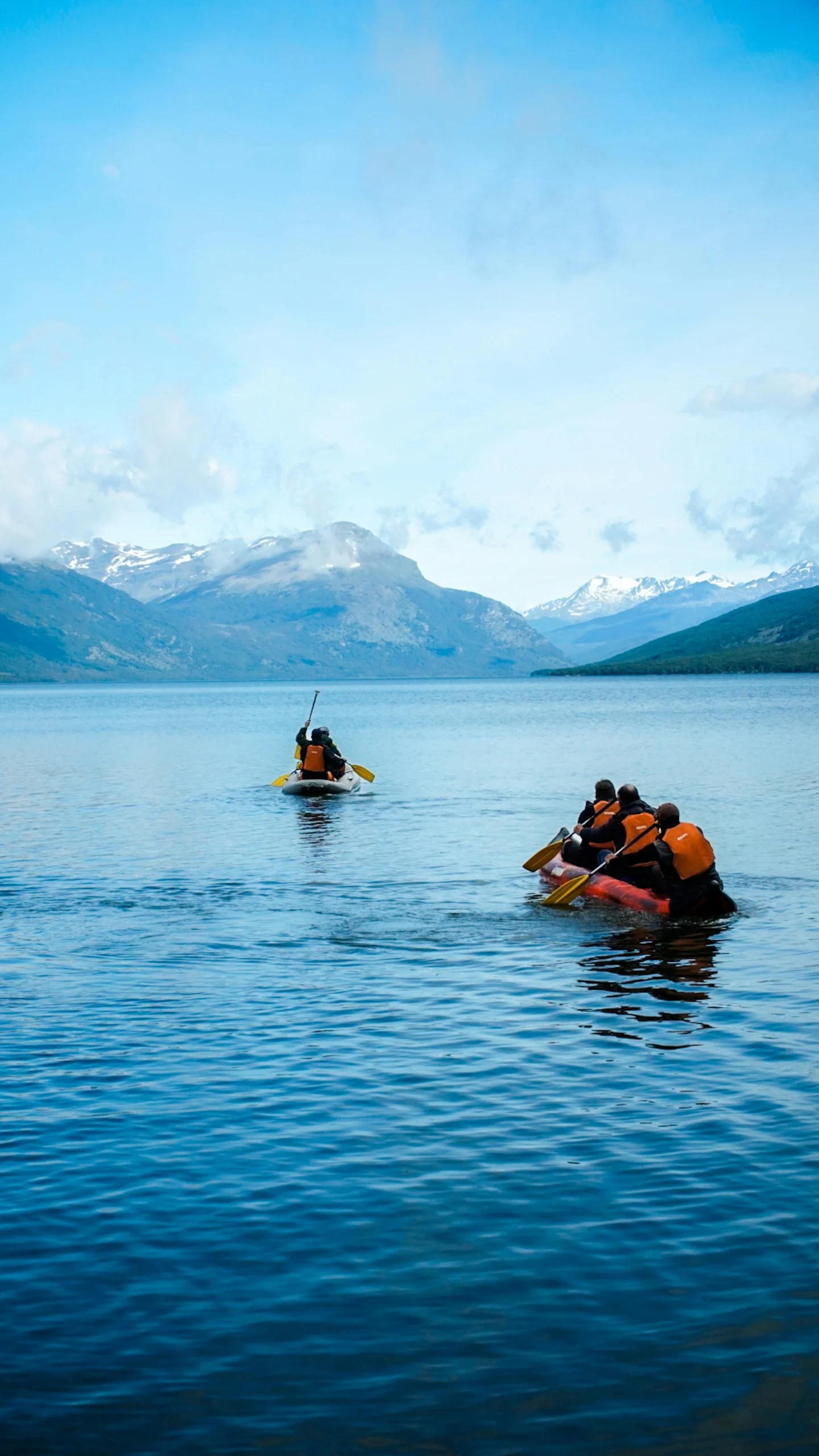A group of people in two canoes on a calm lake with a wide-open sky and a mountain range in the background.
