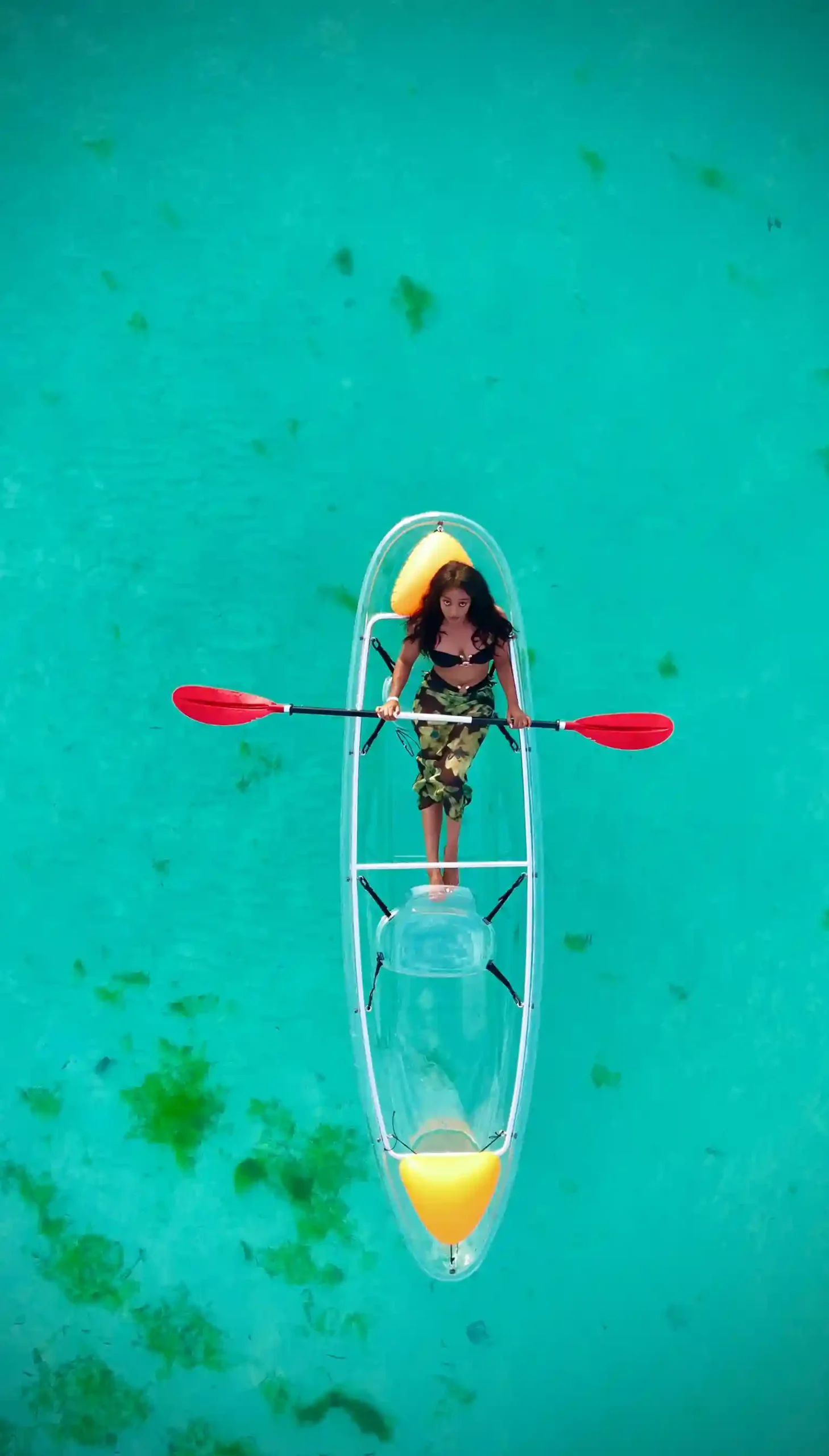 An overhead shot of a person standing in a clear kayak, paddling through the turquoise, shallow waters of Zanzibar.