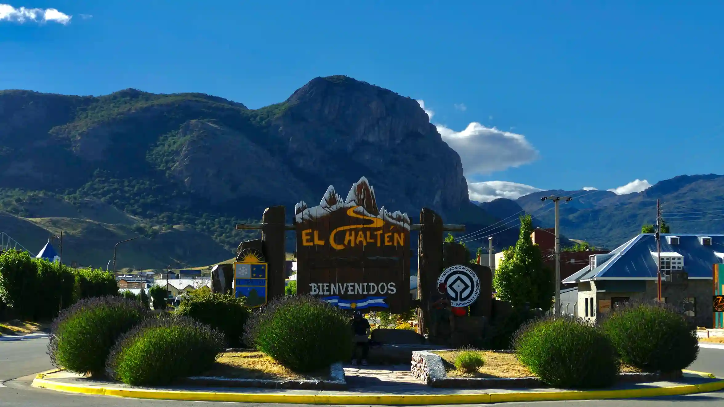 A wooden welcome sign for the town of El Chaltén, with the Andes mountains in the background under a blue sky.