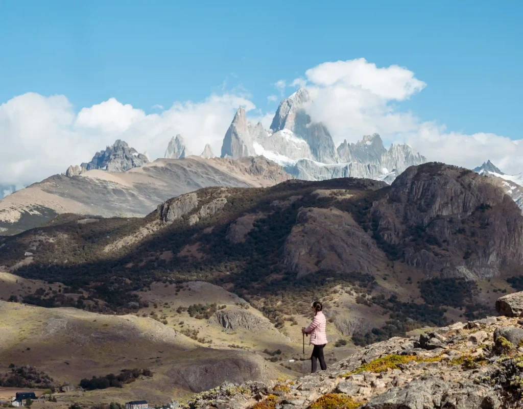A lone hiker with trekking poles stands on a rocky path, admiring the view of the jagged peaks of Mount Fitz Roy under a clear sky.