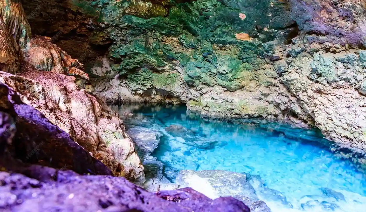 The interior of Kuza Cave in Zanzibar, showing a natural freshwater pool with vivid blue water surrounded by colored rock formations.