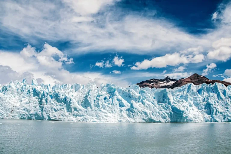 A wide, expansive glacier wall with a rough, icy texture, a mountain visible in the background under a blue sky with scattered clouds.