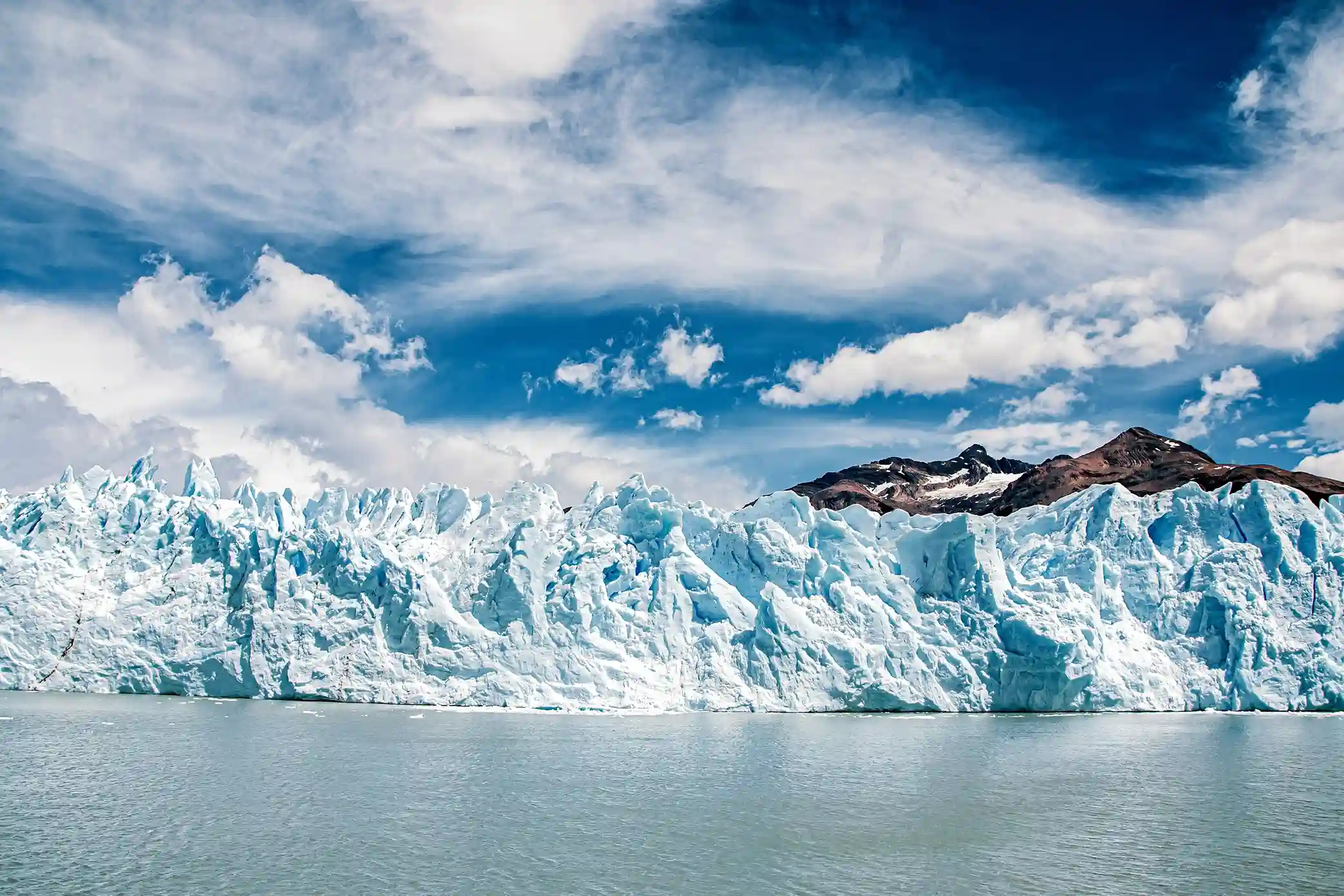 A wide, expansive glacier wall with a rough, icy texture, a mountain visible in the background under a blue sky with scattered clouds.