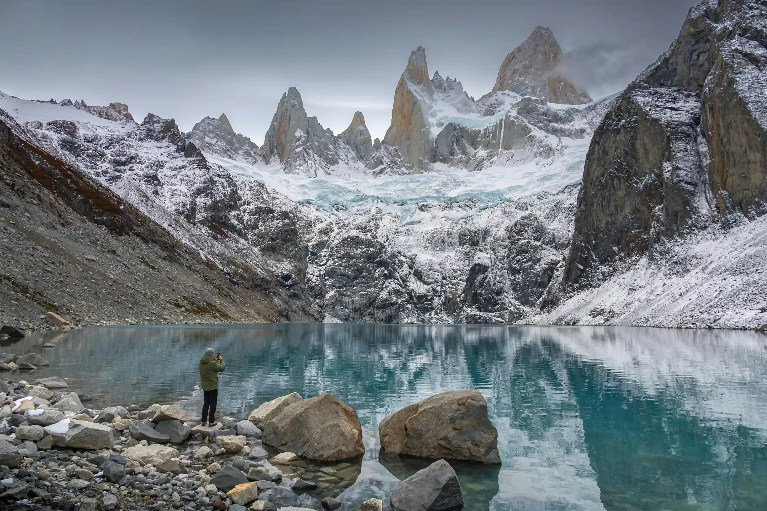 A solitary hiker stands on a rocky shore, looking out at the snow-capped Monte Fitz Roy mountains reflecting in a calm, clear lake.