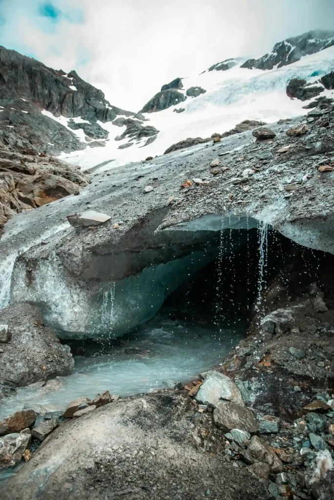 Water drips down from the jagged, translucent blue entrance of a glacier ice cave, with a stream flowing over rocks in the foreground and a snow-capped mountain behind it.