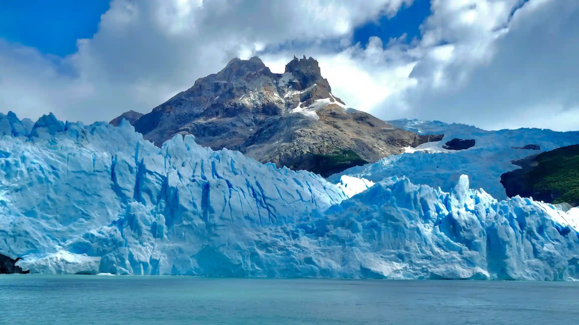 A massive, blue-tinged glacier with a jagged surface sits in front of a dark mountain peak under a partly cloudy sky.