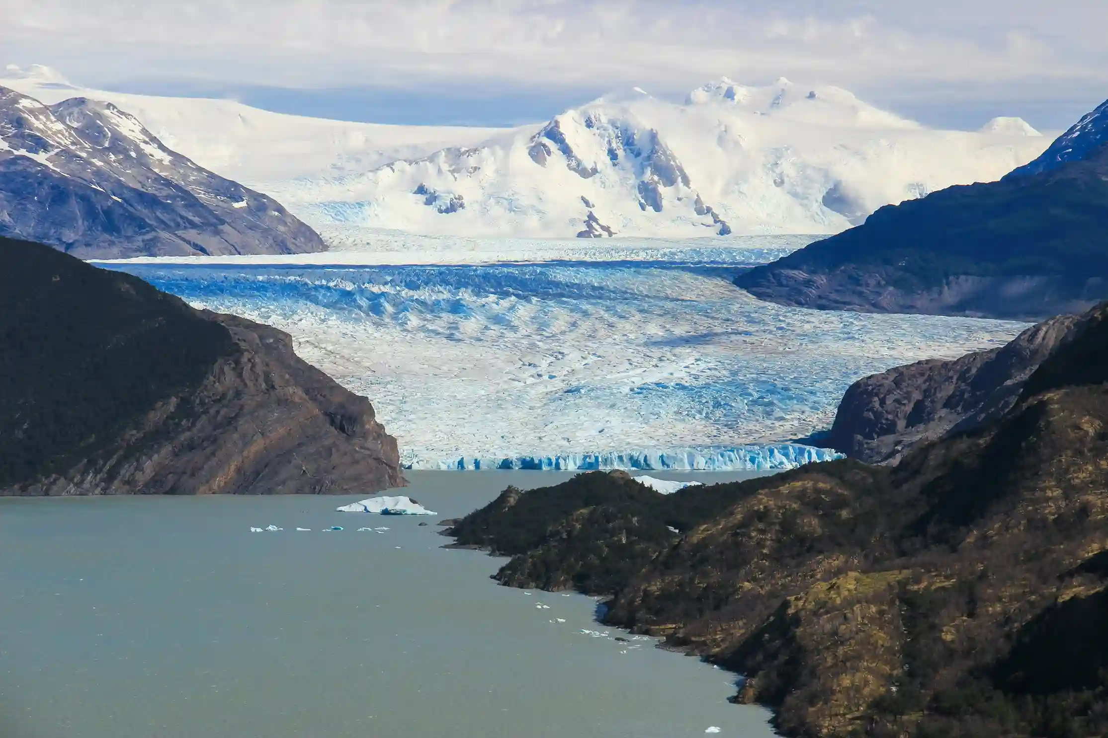 A massive blue and white glacier with a frozen lake at its base, surrounded by a mountain range with snowy peaks.