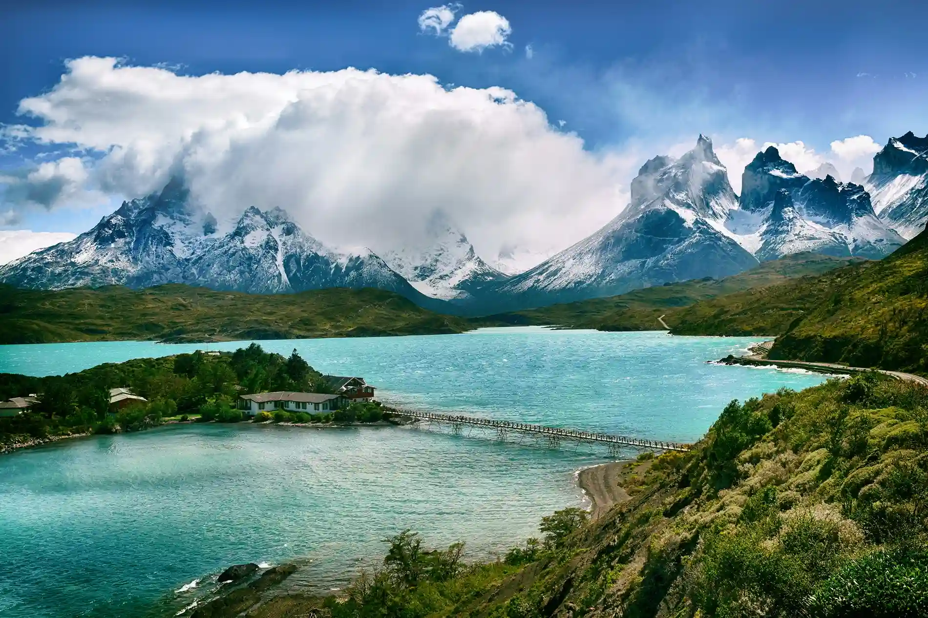Torres del Paine mountains rising behind a turquoise lake with a small wooden bridge and building.