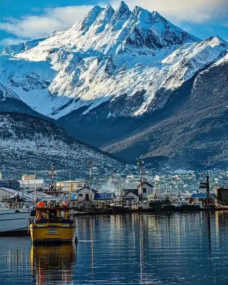 The port of Ushuaia with fishing boats in the foreground and a large, snow-covered mountain rising majestically in the background.