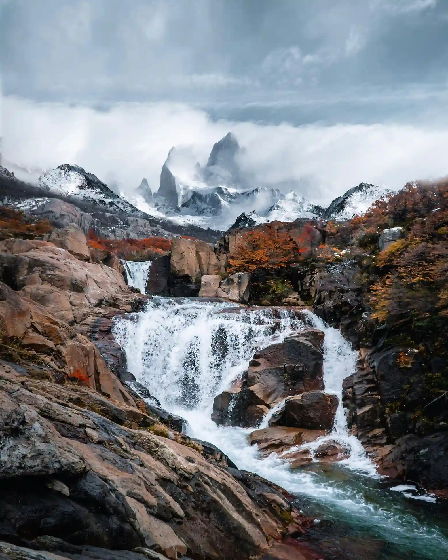 A powerful waterfall cascading over a rocky stream with autumn foliage and the snow-covered peaks of Mount Fitz Roy in the distance.