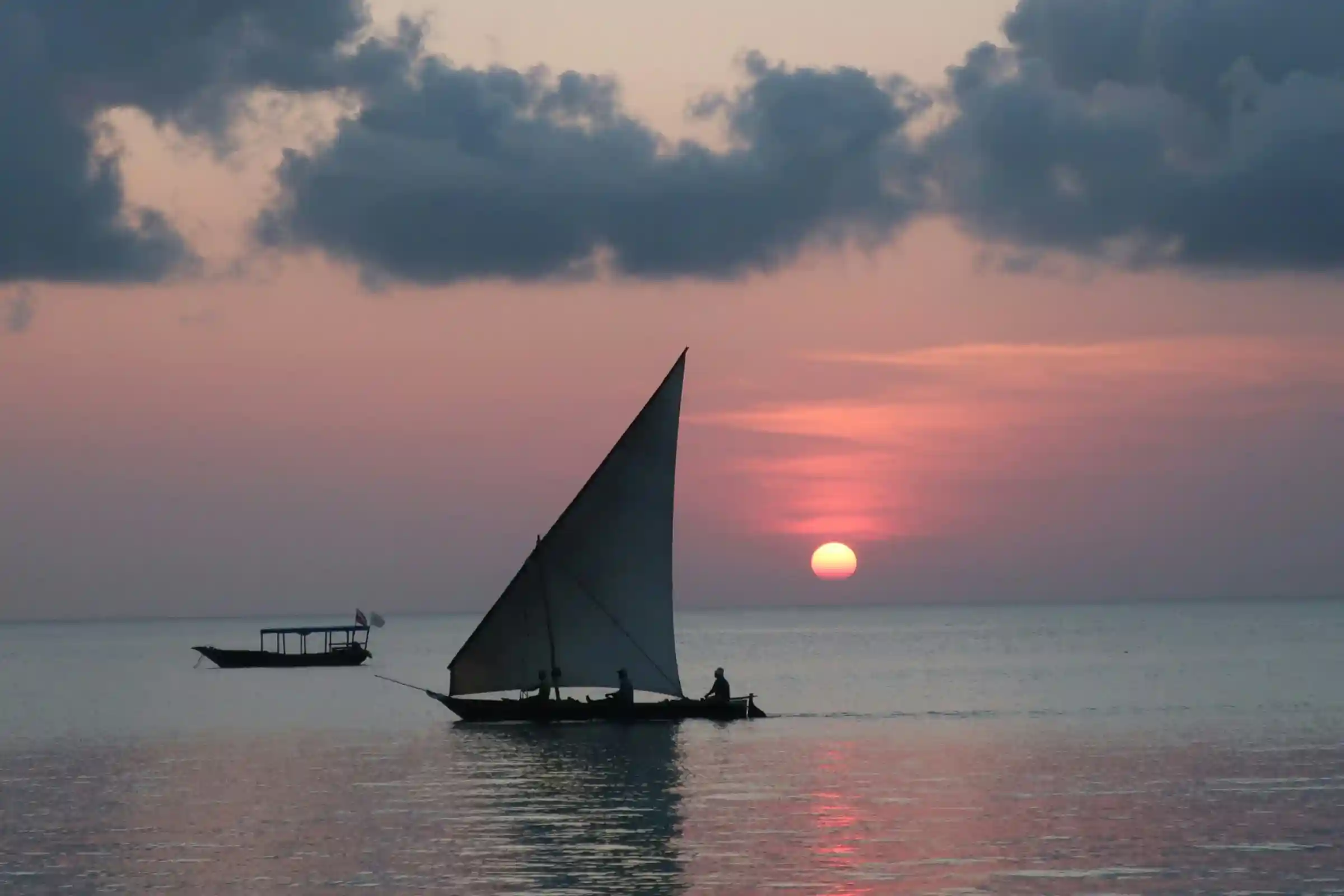 A traditional wooden dhow sailboat silhouetted against a beautiful sunset sky over the Indian Ocean in Zanzibar.