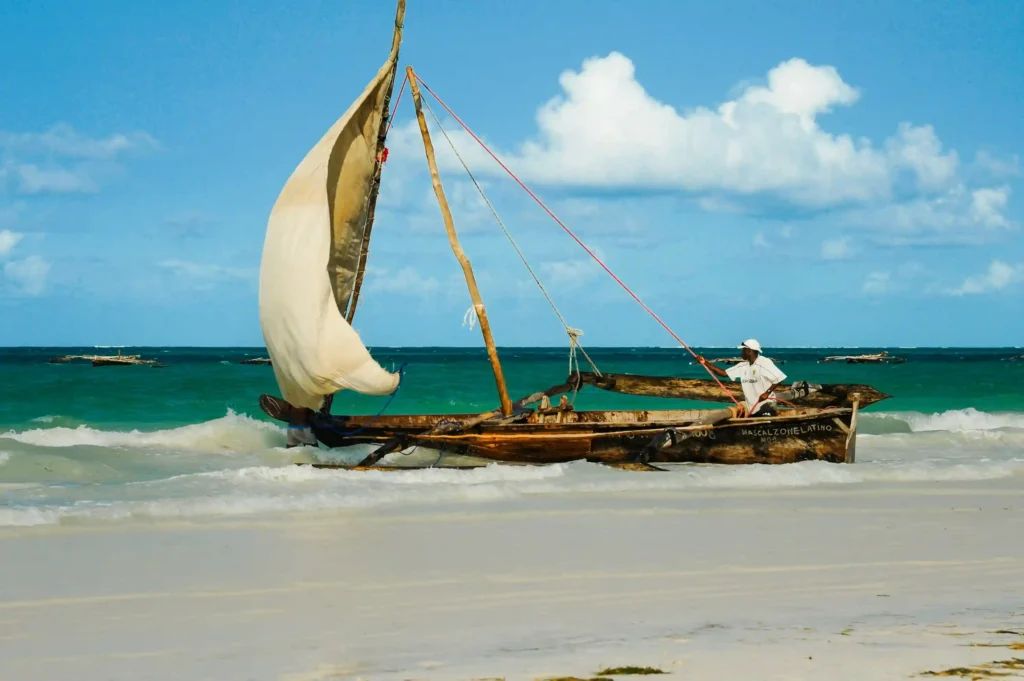 A traditional wooden Zanzibari fishing dhow boat with a white lateen sail is beached on a white sand shore with turquoise water, a fisherman in white clothing sits near the stern.