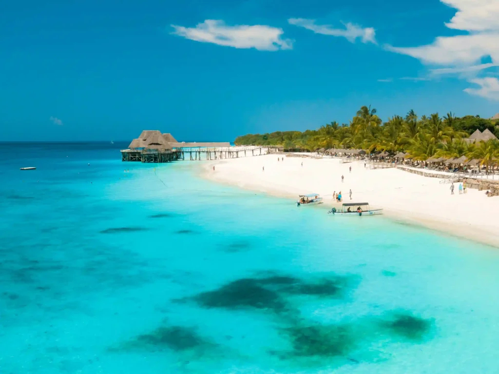Aerial view of a luxury resort on a white sand beach in Zanzibar with vibrant turquoise water, a wooden pier, and palm trees.